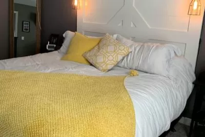 A cozy bedroom featuring a bed with a white modern headboard, a bright yellow knitted throw over white bedding, and industrial-style wall lamps