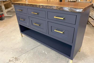 A modern dresser in navy blue with a polished wood countertop, featuring drawers with brass handles and a lower shelf