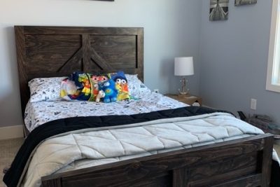 A rustic wooden bed with a high headboard, adorned with colorful plush toys, set against a pale blue wall with a sign reading "CAPTAIN`S QUARTERS" above it