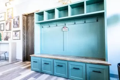 A blue wooden Mudroom bench with matching wall-mounted shelves and coat hooks in a bright room with a hardwood floor.