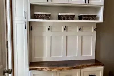 A white mudroom bench with baskets in the middle section above coat hooks and a door 